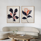 A modern living room featuring a curved beige sofa, a round wooden coffee table with vases and magazines, and the Blush & Shadow Set of 2 framed abstract floral artworks in black and peach tones on the wall.