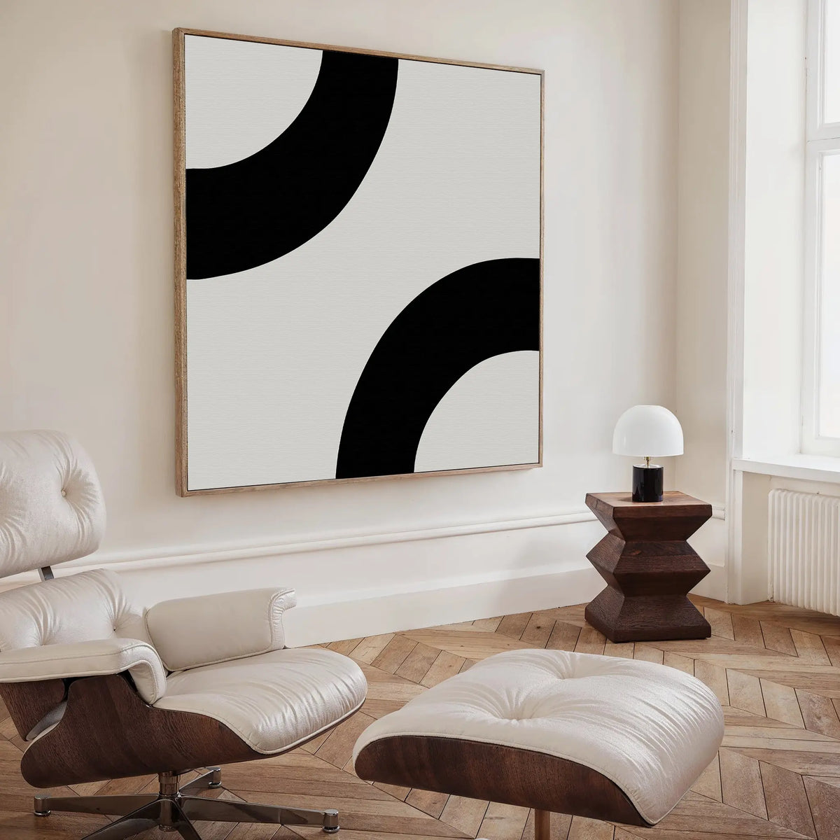 A modern, minimalist living room features a white lounge chair with ottoman, a geometric wood side table with a small lamp, and Bold Orbit Canvas Art on the wall. Light wood floors and a large window enhance the bright, airy space.