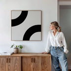 A woman in a white shirt and blue jeans stands by a wooden sideboard, admiring the Bold Orbit Canvas Art on the wall. A potted plant and books enhance the modern decor, creating a stylish yet inviting atmosphere.