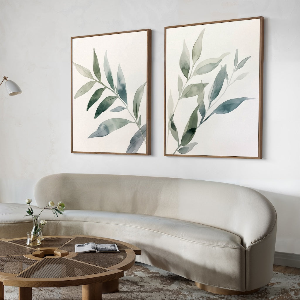 A modern living room with a curved beige sofa, a round wooden coffee table topped with a glass vase and open book, and the Botanical Breeze Set of 2 minimalist wall art prints featuring green leaves displayed above the sofa.