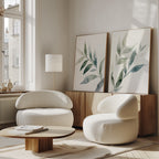 A modern living room features the Botanical Breeze Set of 2 white curved armchairs, a low wooden coffee table, and green leaf minimalist wall art on a light wall near a large window, with natural light brightening the space.
