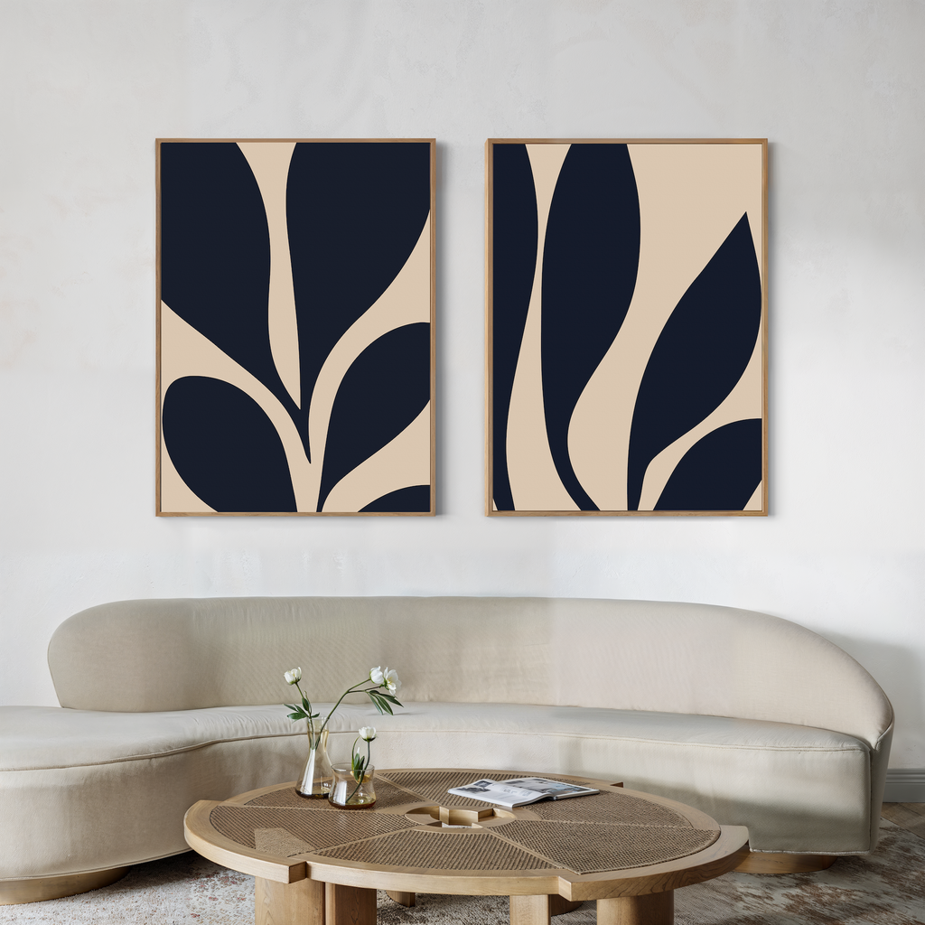 A modern living room with a curved beige sofa, a round wooden coffee table topped with flowers, and the Botanical Ink Set of 2 minimalist wall art in navy and beige adding style to the light wall.