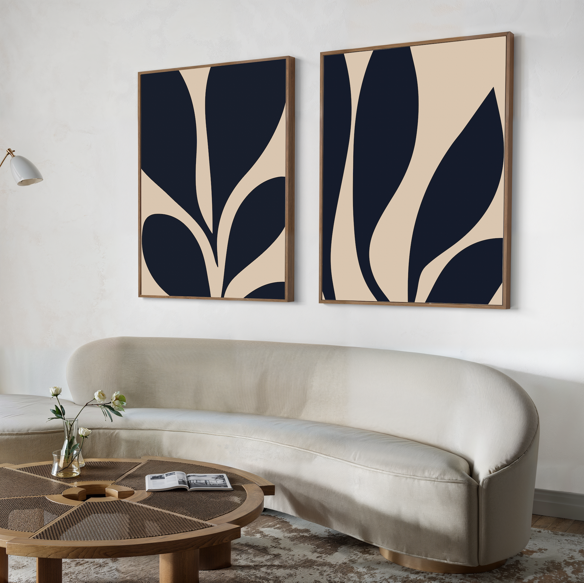 A modern living room features a curved cream sofa, a round wooden coffee table with a vase and books, and the Botanical Ink Set of 2—minimalist blue and beige botanical wall art—elegantly displayed above as decor.