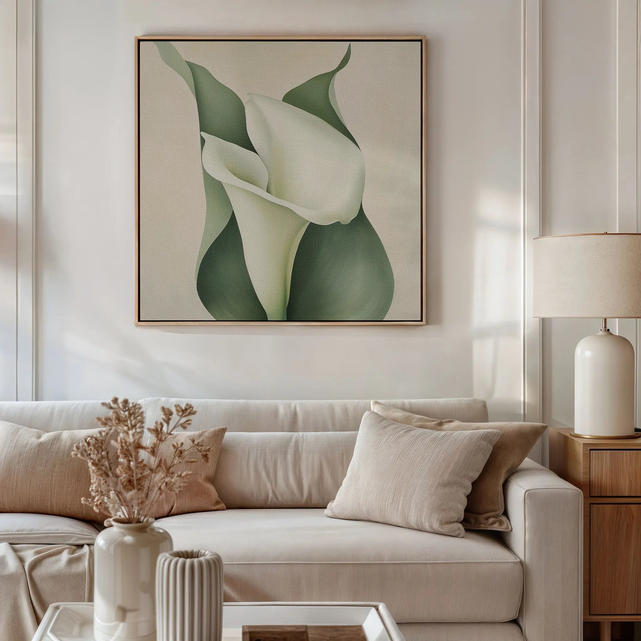 A neutral-toned living room with a light beige sofa, soft pillows, a side table and lamp, dried flowers in a vase, and striking floral wall art—the Calla Duo Canvas Art—featuring white calla lilies with green leaves as part of the modern decor.