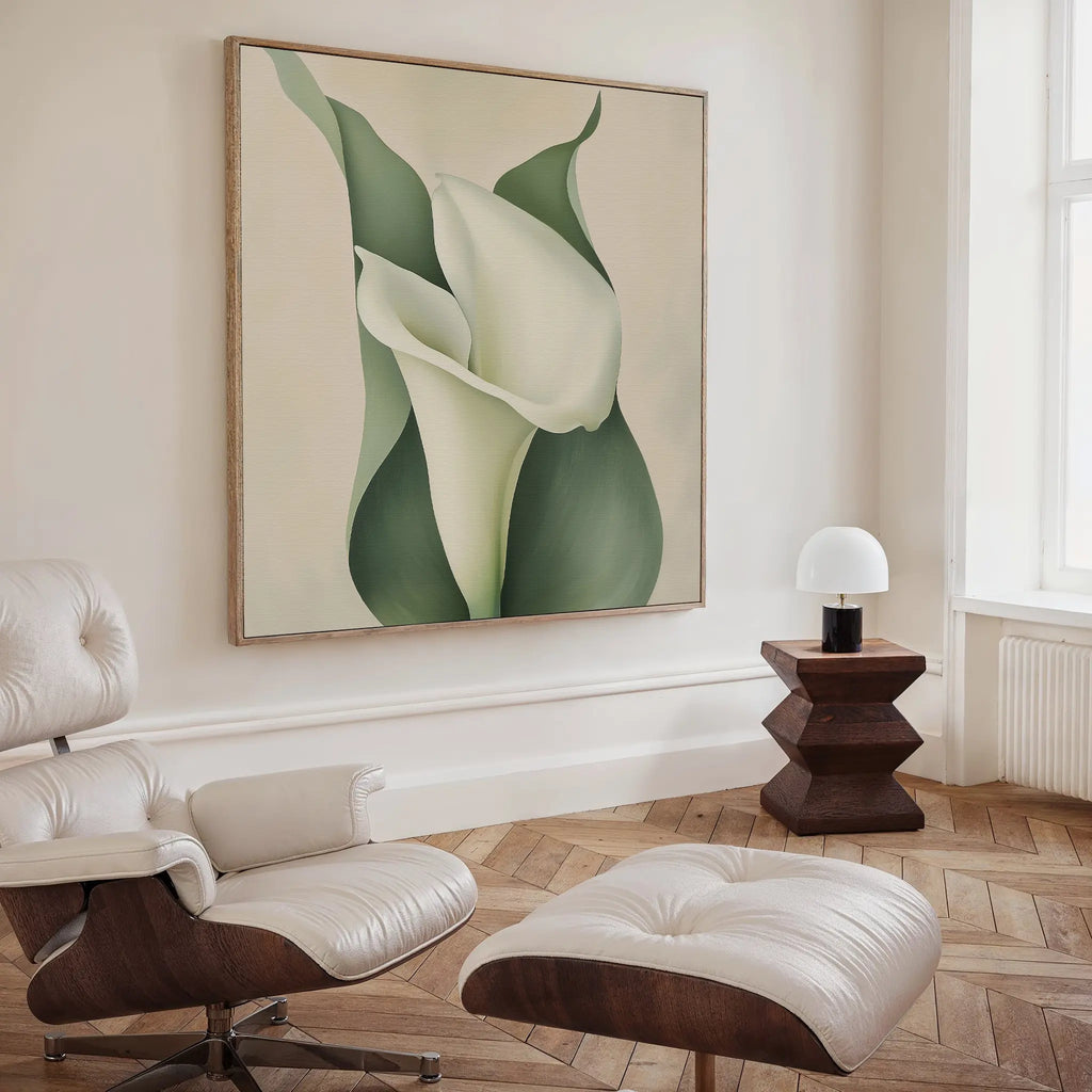 A modern living room with a white leather lounge chair and ottoman, a wooden side table with a white lamp, and the Calla Duo Canvas Art featuring a large white flower with green leaves on the wall.