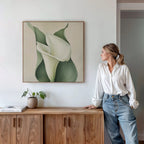 A woman in a white blouse and jeans leans against a wooden cabinet, gazing at the Calla Duo Canvas Art—a minimalist painting of a white calla lily with green leaves. A small potted plant and books add to the modern decor on the cabinet.