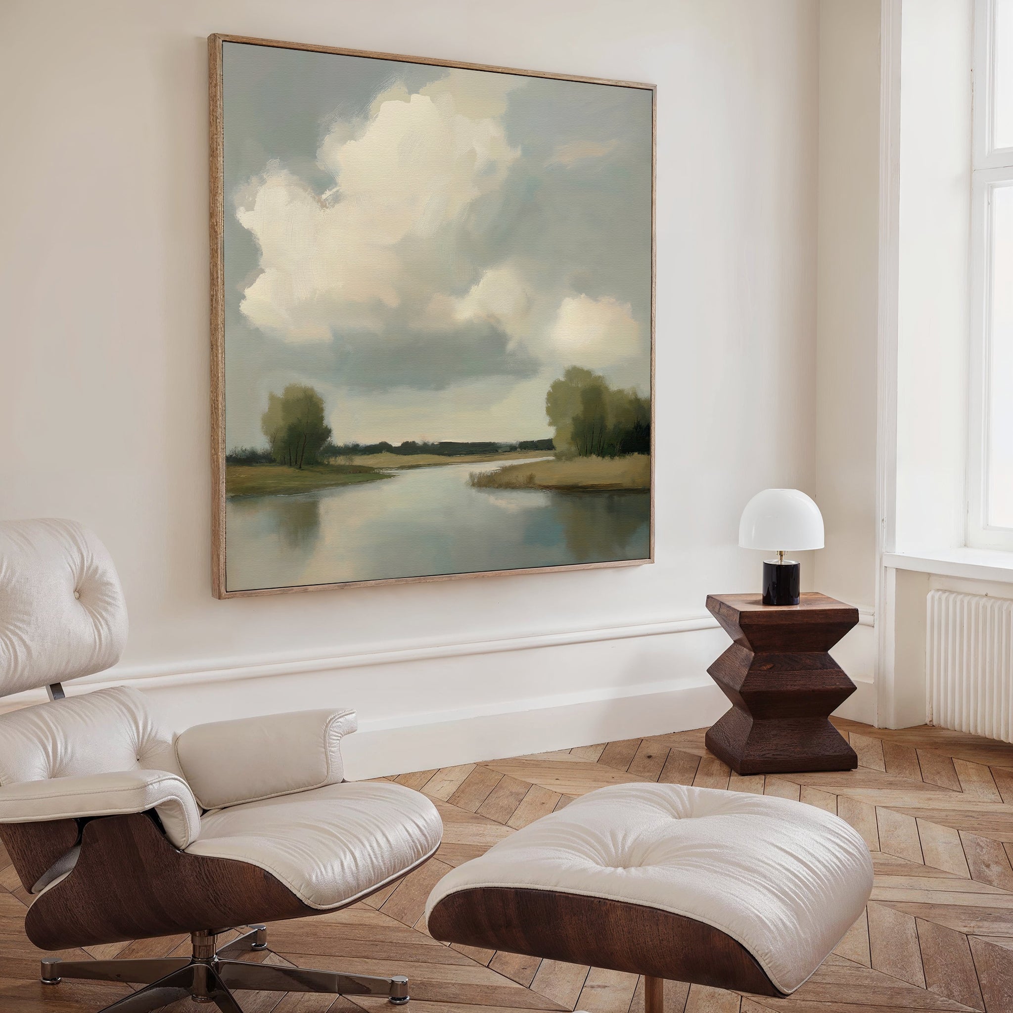 A modern living room featuring a white leather lounge chair and ottoman, a small wooden side table with a white lamp, and the Calm Passage Canvas Art—a large minimalist river-and-cloud painting—creates an inviting, tranquil space.