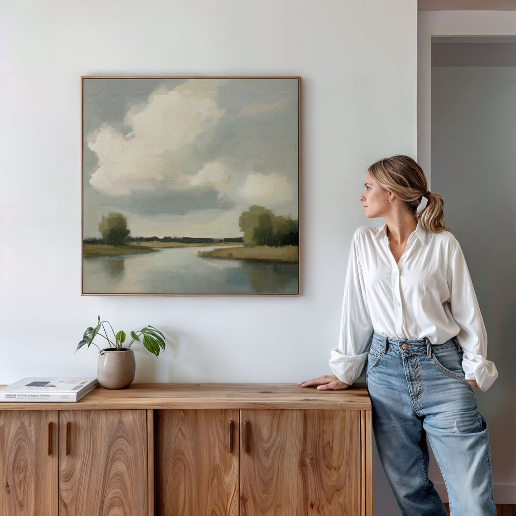 A woman in a white blouse and jeans leans against a wooden cabinet, admiring the Calm Passage Canvas Art—a minimalist landscape painting of a cloudy sky and river—hanging on a white wall. A potted plant and books complete the serene bedroom decor.