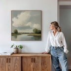 A woman in a white blouse and jeans leans against a wooden cabinet, admiring the Calm Passage Canvas Art—a minimalist landscape painting of a cloudy sky and river—hanging on a white wall. A potted plant and books complete the serene bedroom decor.