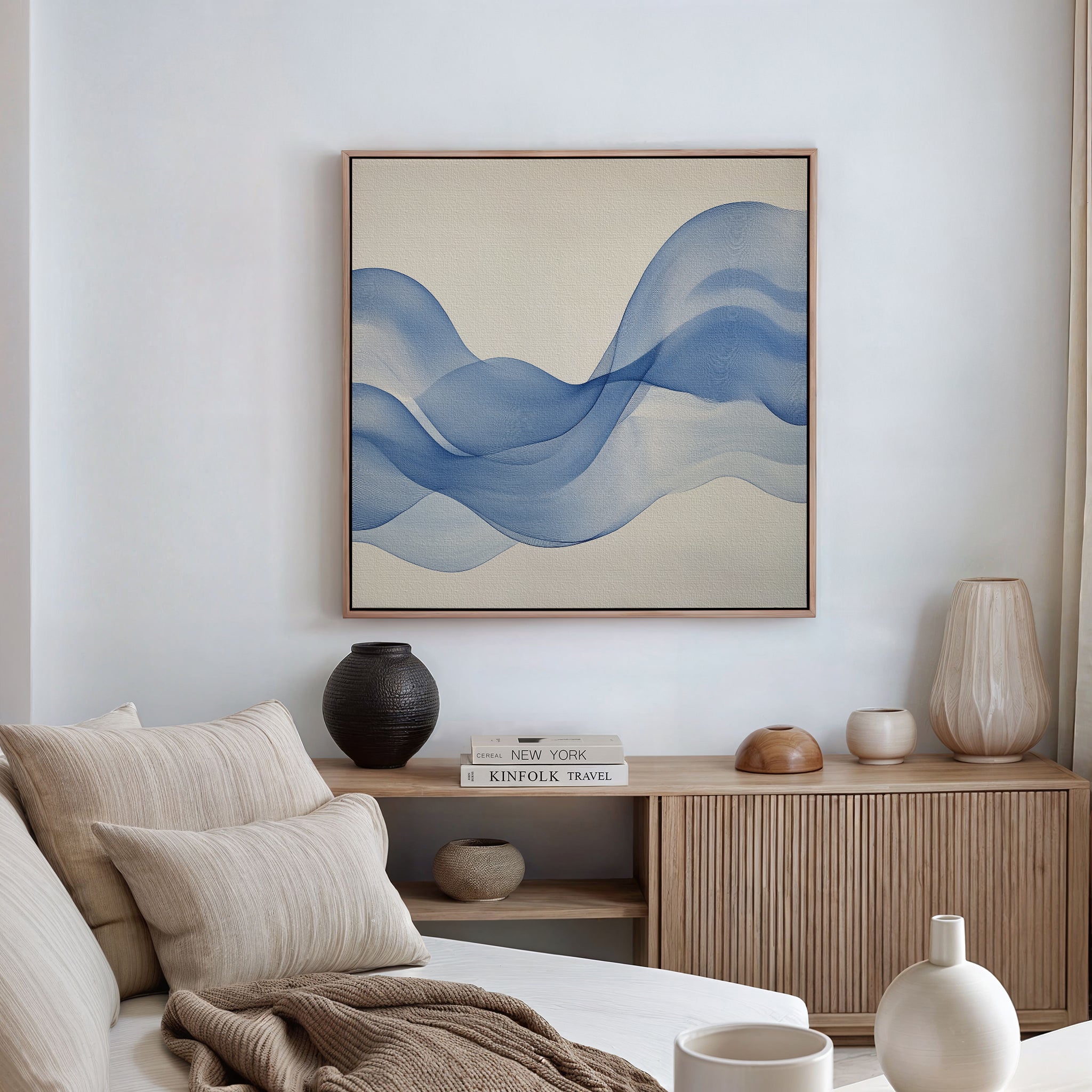 A minimalist living room features a beige sofa, wooden sideboard with vases and books, and the Calm Swell Canvas Art—a large abstract blue waves print. Soft natural light enhances the spaces modern decor.