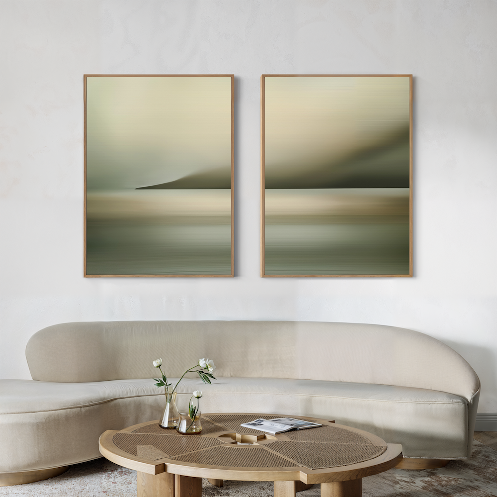 A modern living room features a curved beige sofa, a round wooden coffee table with a vase of flowers, and the Calm Waterscape Set of 2 paintings above the sofa in earthy green and beige tones.