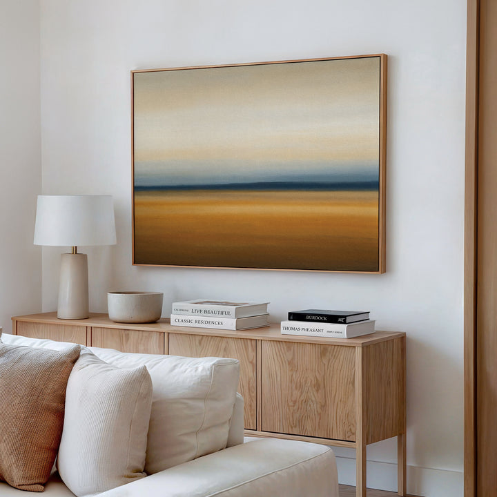 A minimal living room with a beige sofa, light wood sideboard, white lamp, stacked books, and the Caramel Plains Canvas Art – Horizontal featuring soft earthy tones on the wall.