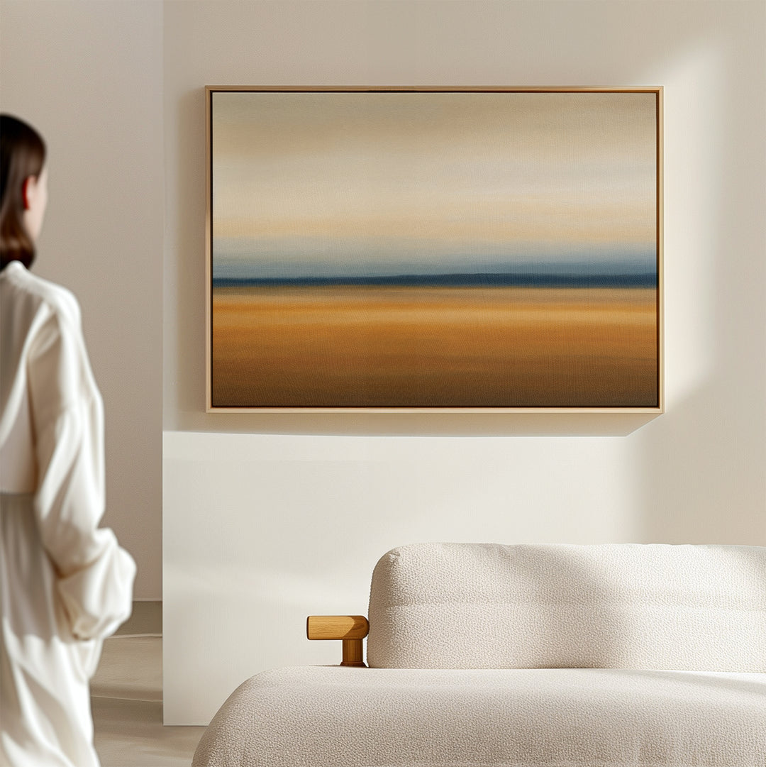 A person in white stands in a minimalist room, admiring the Caramel Plains Canvas Art – Horizontal, featuring abstract beige, blue, and brown bands above a light-colored sofa.