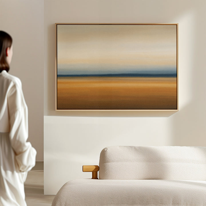 A person in white stands in a minimalist room, admiring the Caramel Plains Canvas Art – Horizontal, featuring abstract beige, blue, and brown bands above a light-colored sofa.