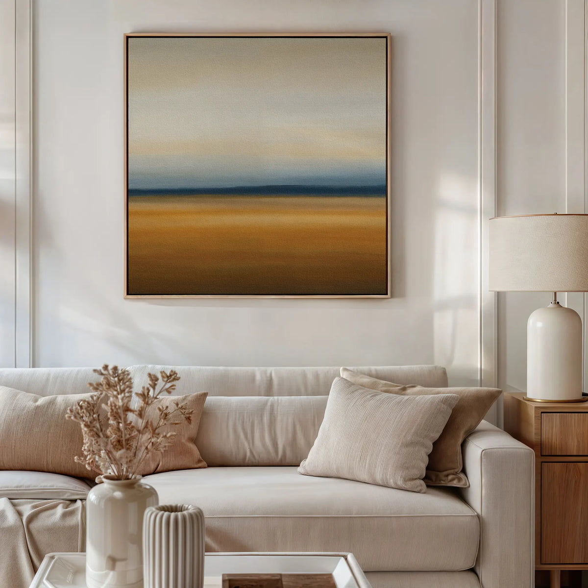 A modern living room with a beige sofa, neutral pillows, a side table with a white lamp, and Caramel Plains Canvas Art in earth tones and blue shades. A vase with dried flowers on the coffee table complements the minimalist decor.