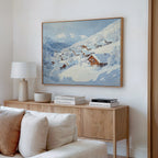 A cozy living room features a light wood sideboard with stacked books, a white lamp, and the Chalet View Canvas Art—a snowy winter village scene—above soft neutral decor and a white sofa in the foreground.