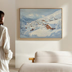 A person stands in a minimal, modern living room, admiring Chalet View Canvas Art—a framed painting depicting a peaceful winter village scene with houses and trees under a bright sky.