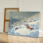 The Chalet View Canvas Art, featuring a winter village with snowy cabins and trees, rests against a white wall on a herringbone floor—perfect minimalist wall decor. A blank canvas leans behind it.