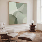 A modern living room featuring a white leather lounge chair and ottoman, a wooden side table with lamp, and the Circular Quiet Abstract Canvas Art in green and beige tones displayed on a cream-colored wall.