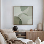 Minimalist living room featuring a cream sofa, neutral cushions, wooden sideboard, vases, books, and modern wall art: the Circular Quiet Abstract Canvas Art in green and beige on a white wall. Natural light streams in from the right.