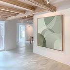 A minimal, modern room with exposed wooden ceiling beams and white walls features the Circular Quiet Abstract Canvas Art—its green and beige overlapping shapes evoke Nordic Park serenity as natural light streams through nearby windows.