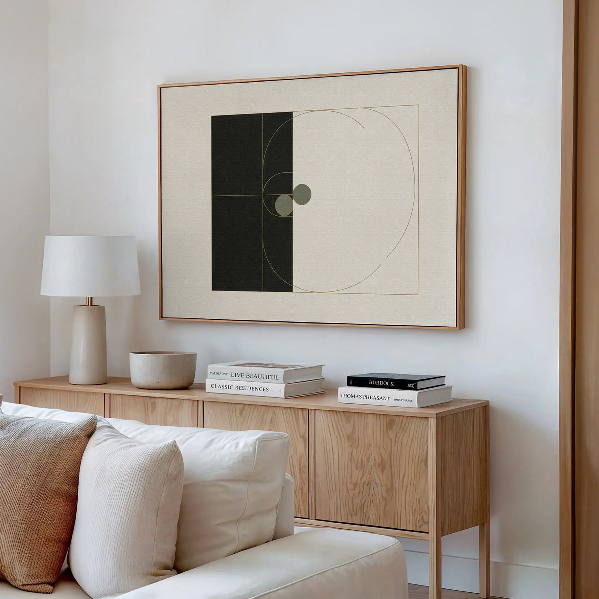 A modern living room features a light wood sideboard with books, a lamp, and a bowl. Above it hangs the Circular Tension Canvas Art with abstract geometric shapes. A white sofa sits in the foreground against a neutral wall.