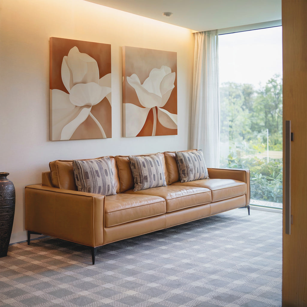 A modern living room with a tan leather sofa, four patterned cushions, a dark vase, sheer curtains on a large window overlooking greenery, and the Clay Blossom Set of 2 floral wall art prints displayed above.