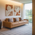 A modern living room with a tan leather sofa, four patterned cushions, a dark vase, sheer curtains on a large window overlooking greenery, and the Clay Blossom Set of 2 floral wall art prints displayed above.