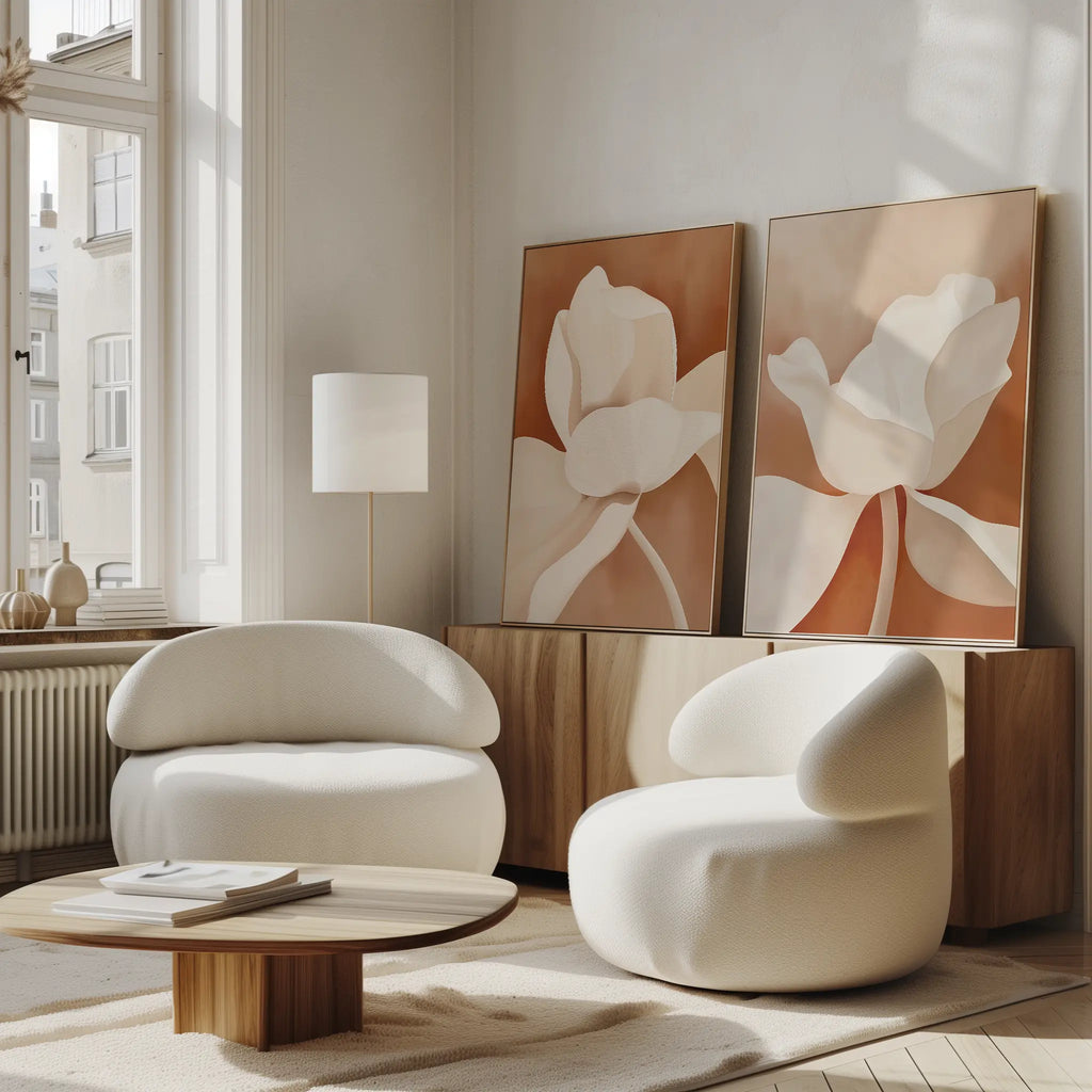 A modern living room with the Clay Blossom Set of 2 rounded white armchairs, a low wooden coffee table, and neutral-toned floral wall art on a wooden sideboard, bathed in sunlight from tall windows.
