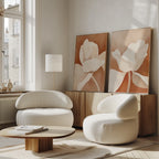 A modern living room with the Clay Blossom Set of 2 rounded white armchairs, a low wooden coffee table, and neutral-toned floral wall art on a wooden sideboard, bathed in sunlight from tall windows.