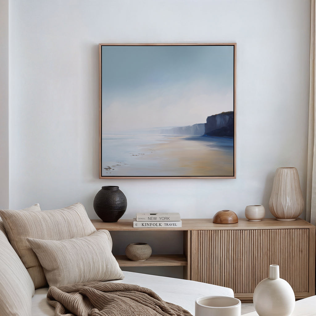 A cozy living room with a beige sofa, neutral cushions, a wooden sideboard topped with vases and books, and large wall art—the Cliffside Light Canvas Art—adds elegance with its serene cliffside scenery.
