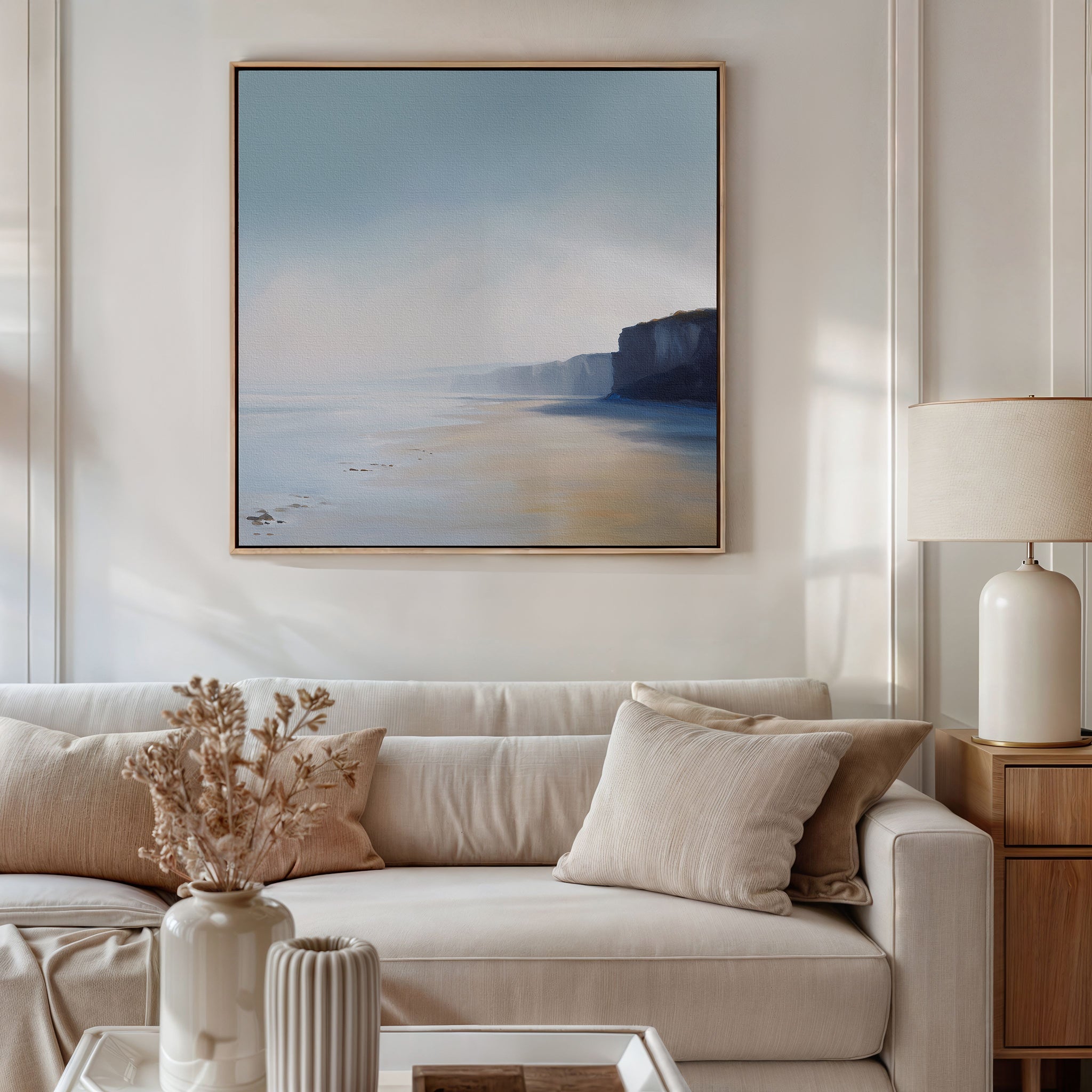 A cozy living room features a beige sofa with neutral cushions, a side table holding a lamp and a vase of dried flowers, and Cliffside Light Canvas Art depicting calm beaches and cliffs under a soft blue sky hanging above the sofa.