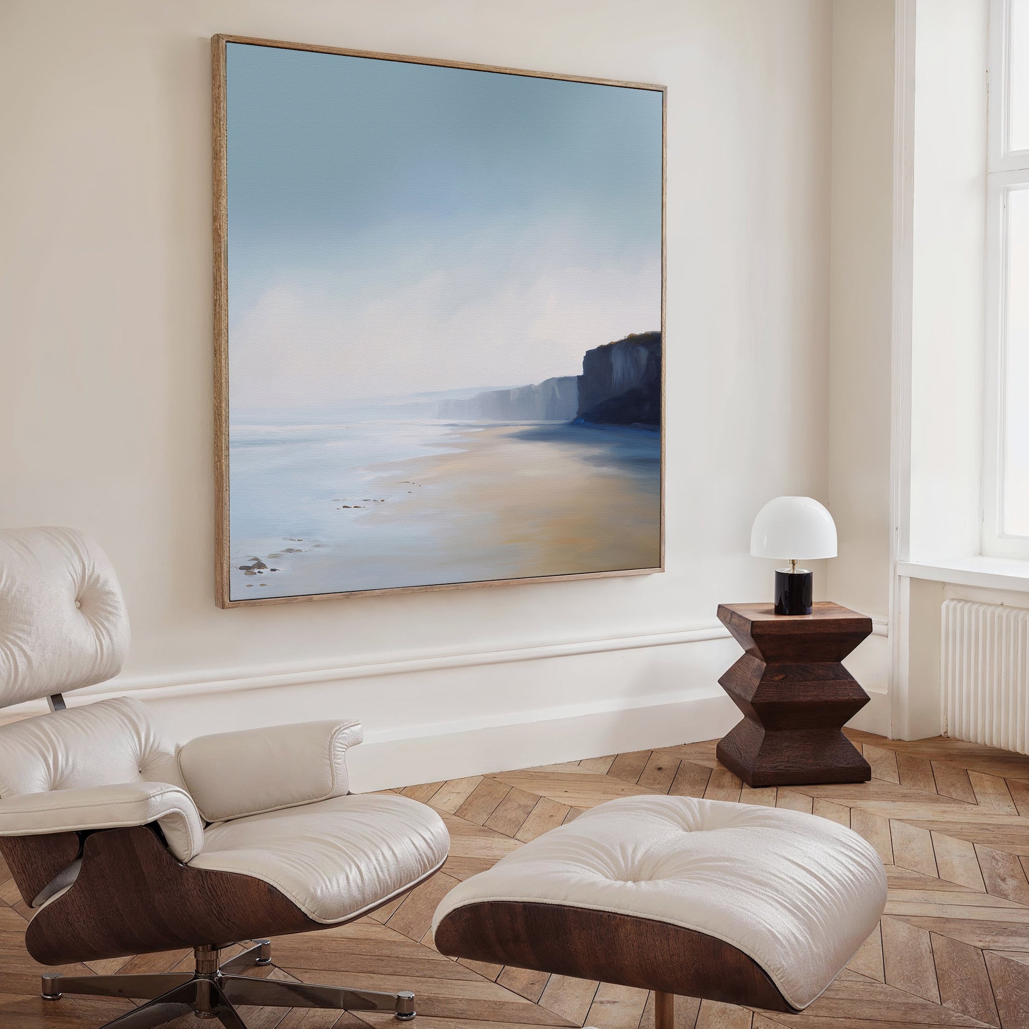 A modern, minimalist living room with a cream lounge chair and ottoman, a wooden side table topped with a white lamp, and striking minimalist wall art—Cliffside Light Canvas Art—framed and hanging on the wall.