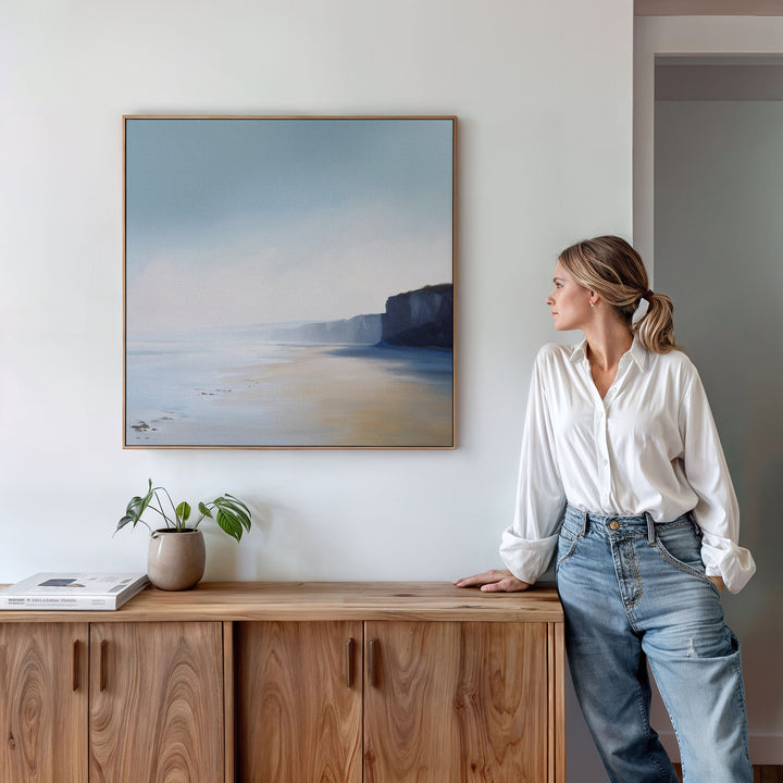 In a bright, modern room, a woman in a white blouse and jeans leans on a wooden cabinet topped with books and a plant, admiring the Cliffside Light Canvas Art displayed above her as part of the stylish, minimalist décor.