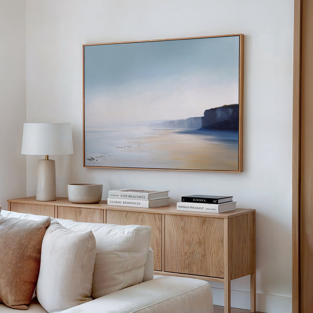 A modern minimalist living room features a wood sideboard, white lamp, stacked books, bowl, and Cliffside Light Canvas Art – Horizontal framed above a beige sofa on a white wall.