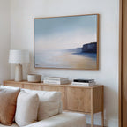 A modern minimalist living room features a wood sideboard, white lamp, stacked books, bowl, and Cliffside Light Canvas Art – Horizontal framed above a beige sofa on a white wall.