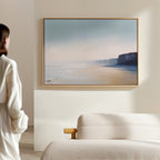 A person in a white shirt stands in a minimalist room, admiring the Cliffside Light Canvas Art – Horizontal, featuring a serene coastal landscape with cliffs and calm sea, framed and mounted above a light-colored sofa.