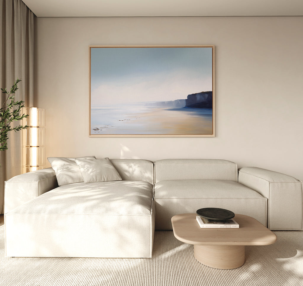 A modern living room with a light beige sectional sofa, wooden coffee table with books and a bowl, leafy plant in the corner, and Cliffside Light Canvas Art – Horizontal above. Natural light fills this inviting space.