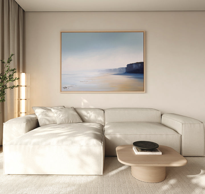 A modern living room with a light beige sectional sofa, wooden coffee table with books and a bowl, leafy plant in the corner, and Cliffside Light Canvas Art – Horizontal above. Natural light fills this inviting space.