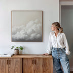 A woman in a white blouse and jeans stands by a wooden cabinet, admiring Cloud Reverie Canvas Art—a minimalist framed painting of fluffy clouds. A potted plant and book on the cabinet complete the calming bedroom decor.
