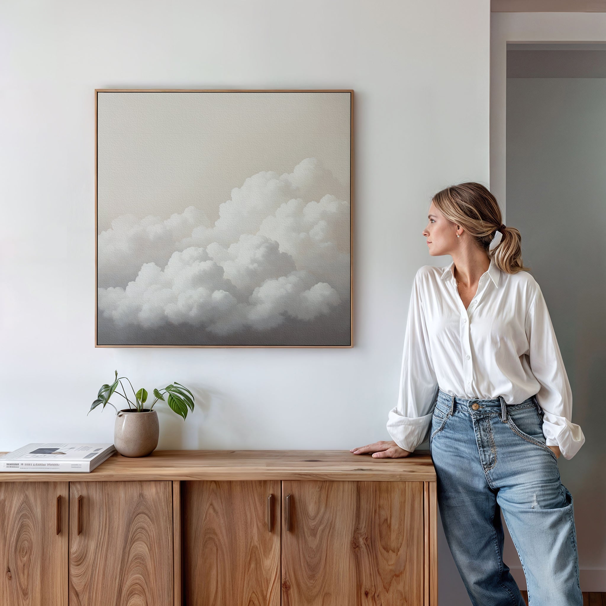A woman in a white blouse and jeans stands by a wooden cabinet, admiring Cloud Reverie Canvas Art—a minimalist framed painting of fluffy clouds. A potted plant and book on the cabinet complete the calming bedroom decor.
