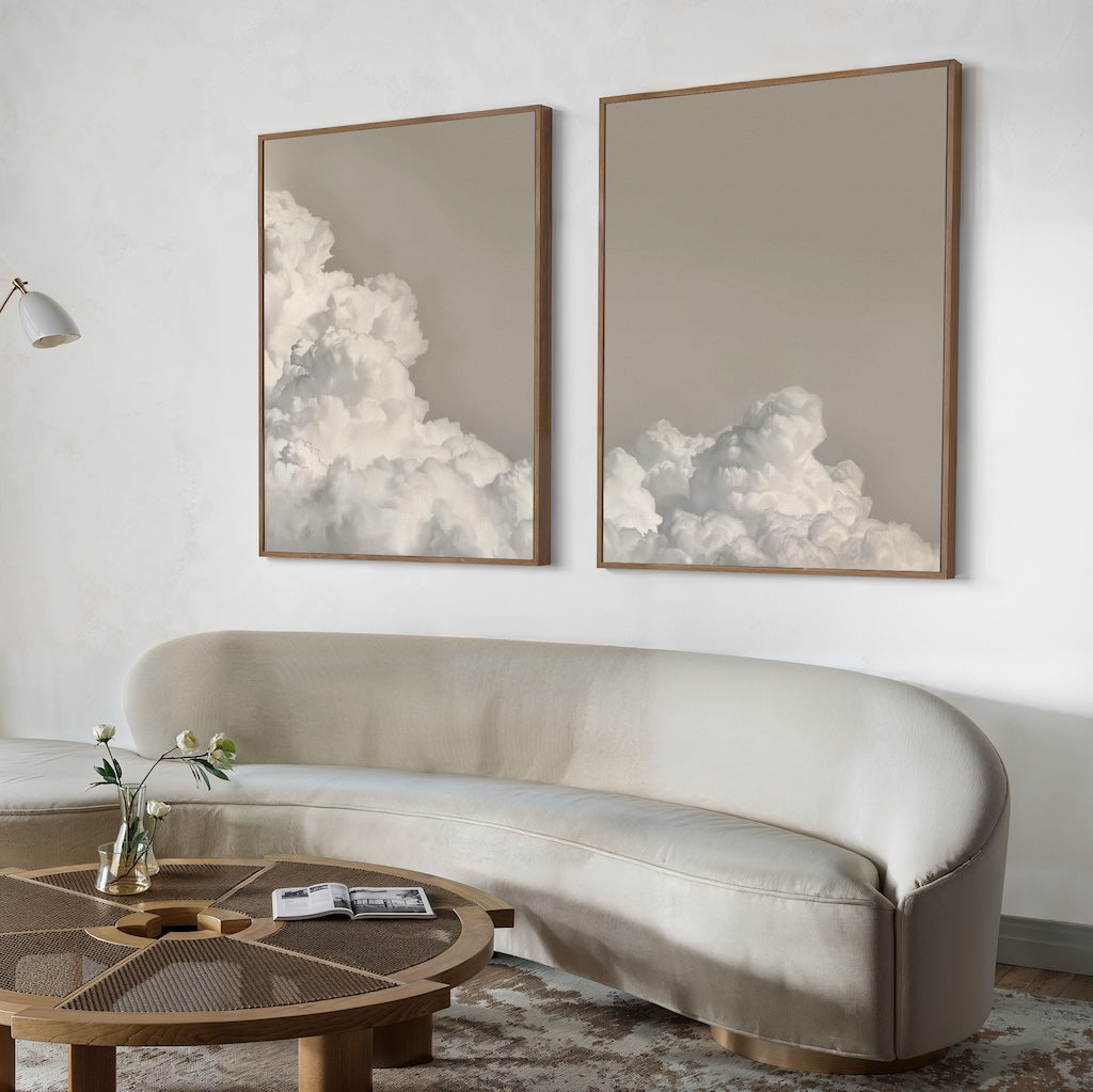 A modern living room with a curved beige sofa, round wooden coffee table, and the Clouded Dreams Set of 2 wall art prints above the sofa. A small vase with flowers and an open book on the table complete this stylish decor.