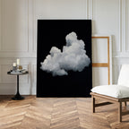 The Cloud In Darkness Abstract Canvas Art, depicting a fluffy white cloud on a black background, is displayed against a white wall in a modern room with herringbone wood floors, a round table with candles and books, and a white cushioned chair.