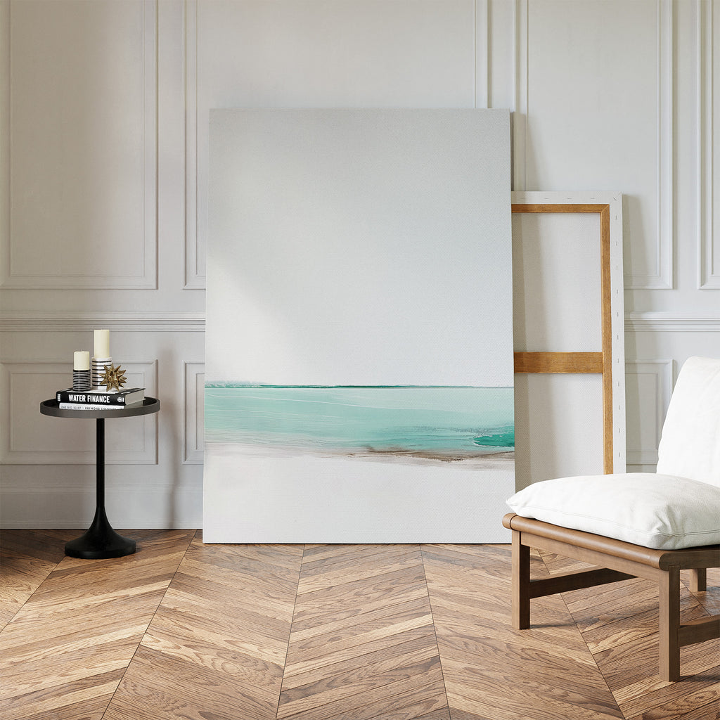 A minimalist room with herringbone wood floors, a white cushioned chair, a black side table with candles and books, and the Coastal Breeze Abstract Canvas Art leaning against a white paneled wall completes the living room decor.