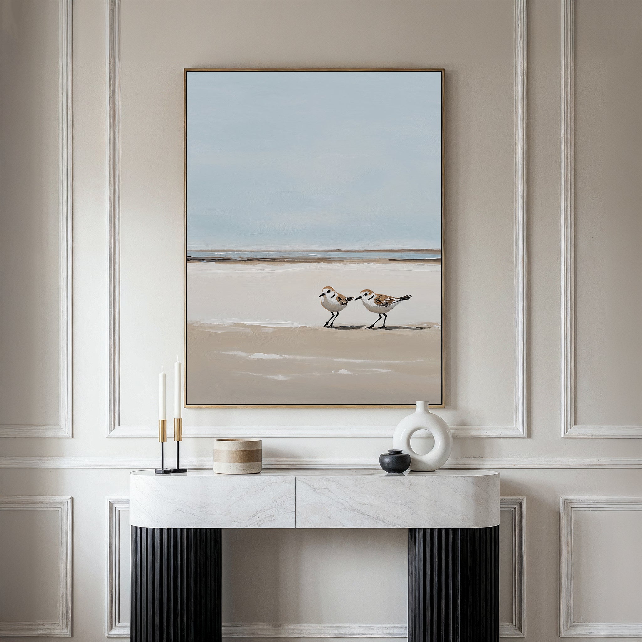 A minimalist console table with decorative objects sits against a white paneled wall, above which hangs the Coastal Chase Canvas Art depicting two shorebirds walking on a beach under a pale blue sky.