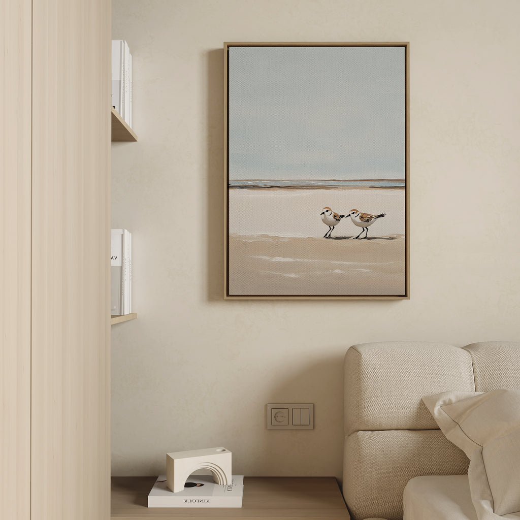 A minimalist living space with a beige wall, cream sofa, wooden bookshelf, and the Coastal Chase Canvas Art depicting two shorebirds on a sandy beach hanging above the sofa.