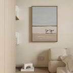 A minimalist living space with a beige wall, cream sofa, wooden bookshelf, and the Coastal Chase Canvas Art depicting two shorebirds on a sandy beach hanging above the sofa.