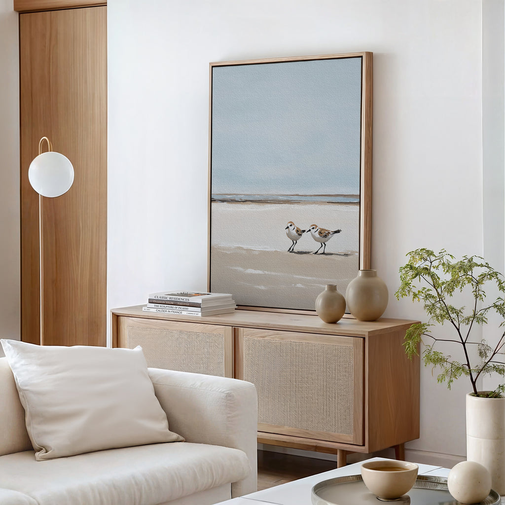 A minimalist living room with neutral tones features a wooden sideboard, vases, books, the Coastal Chase Canvas Art, a cream sofa with a pillow, and a small plant on a round coffee table.