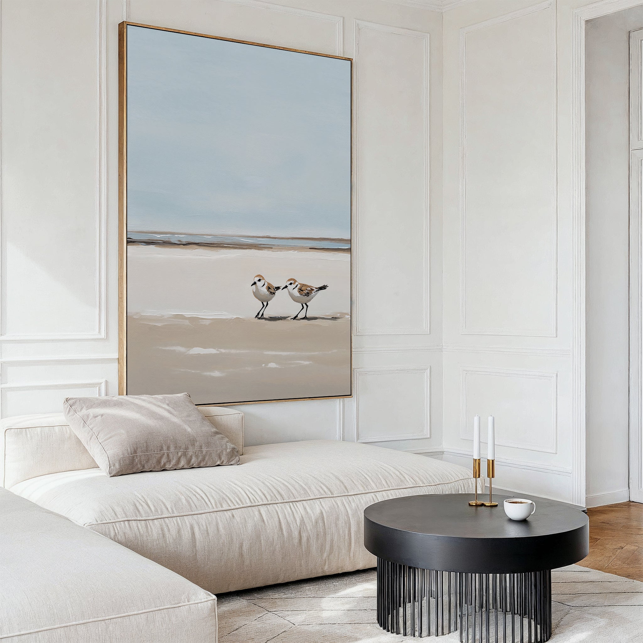 A minimalist living room with a cream sectional, beige pillow, and round black coffee table features the Coastal Chase Canvas Art—a large wall print of two shorebirds—against white paneled walls and warm wood flooring.