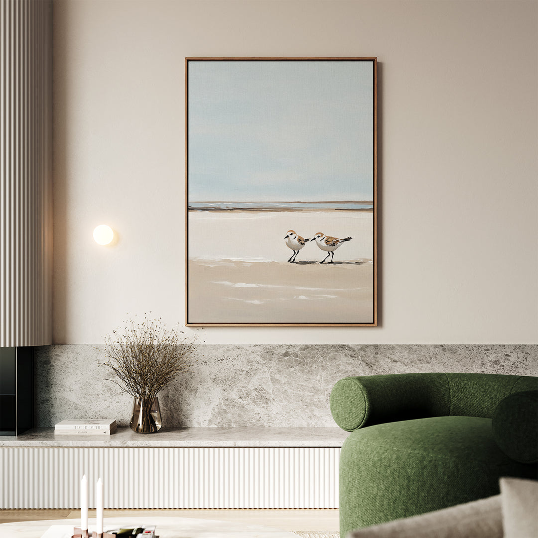 A minimalist living room featuring a green sofa, wall-mounted light, and the Coastal Chase Canvas Art depicting two shorebirds on sand, hanging above a marble shelf with dried flowers and stacked books.